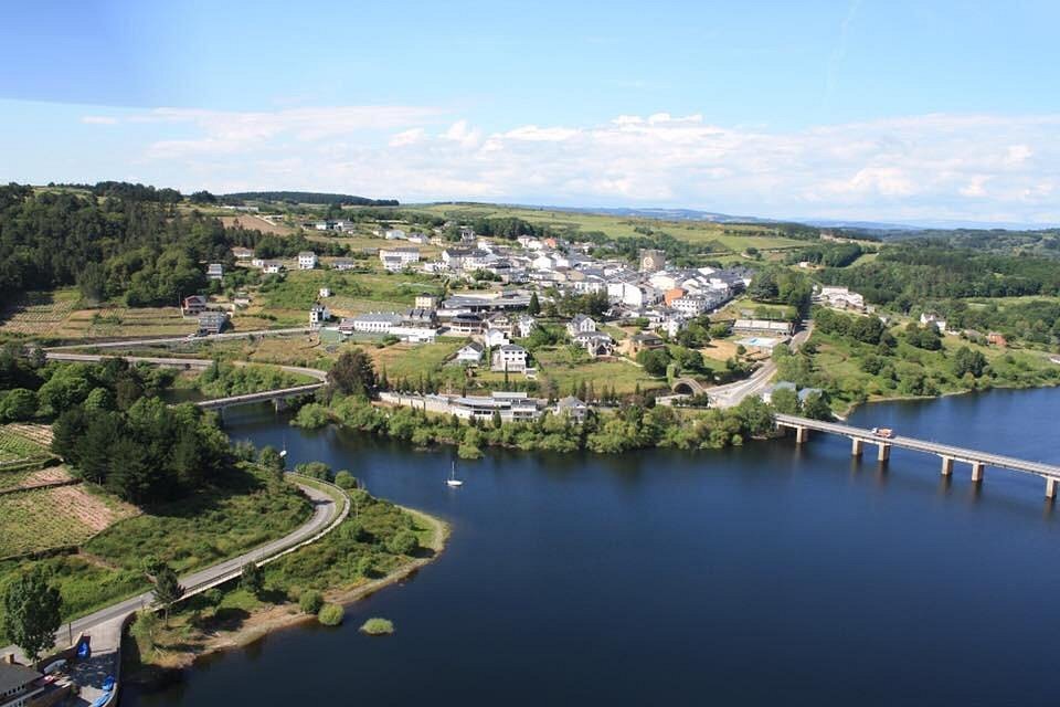 Vista aérea del pueblo de Portomarín en Galicia, a orillas del Embalse de Belesar, con sus casas blancas, colinas verdes y dos puentes que cruzan el río Miño en un día despejado.